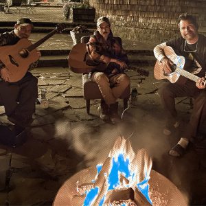 Three guitarists playing acoustic guitars around a firepit