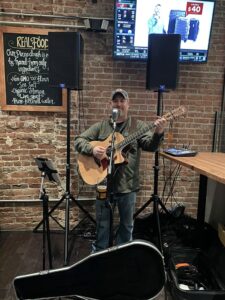 Jeremiah Williams singing and playing acoustic guitar in front of a brick wall