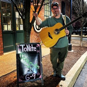 Jeremiah Williams with an acoustic guitar beside a chalkboard announcing his performance