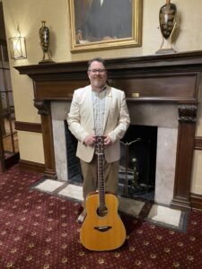 Jeremiah Williams in a cream jacket with an acoustic guitar in front of a fireplace